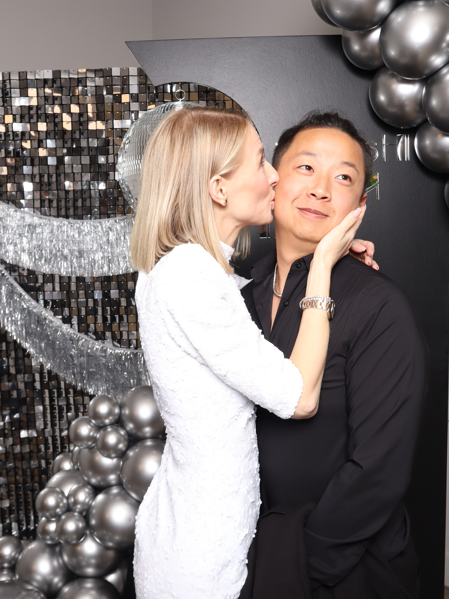 A woman in a white dress kissing a man on the cheek in front of silver balloons and tinsel